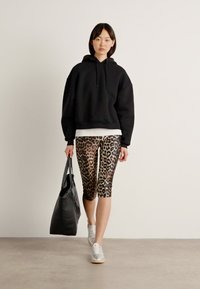 Zwarte oversized hoodie, witte shirt eronder, legging met panterprint tot de kuit, grijze sneakers en een zwarte schoudertas. Soepele texturen.