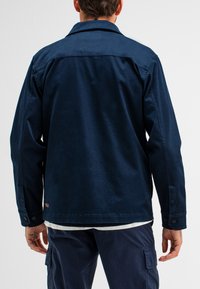 Dunkelblaue Canvas-Jacke mit einem geknöpften Ärmel und klassischem Kragen. Verfügt über eine glatte Textur und ein dezentes Logo nahe dem Saum.