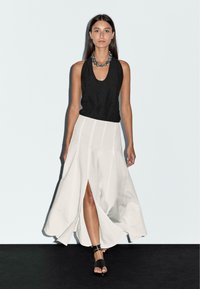 Schwarzes ärmelloses Oberteil, kombiniert mit einem weißen, fließenden, knöchellangen Rock mit einem Schlitz vorne. Schwarze Sandalen mit Riemen runden das Outfit ab.