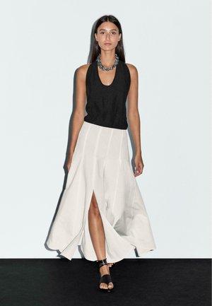 Haut sans manches noir associé à une jupe blanche fluide, longueur cheville, avec une fente à l'avant. Des sandales noires à brides complètent l'ensemble.