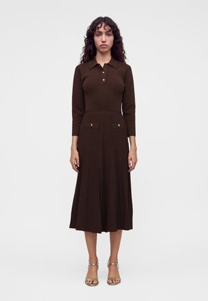Femme aux cheveux bouclés foncés portant une robe marron à manches longues avec des boutons dorés et une jupe plissée, debout devant un fond uni.