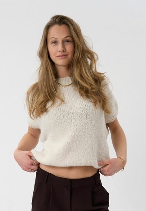 Noor naine pikkade blondide juustega, kes kannab valget lühikeste varrukatega kootud kampsunit ja tumedaid pükse, hoides kampsuni ääre mõlema käega.