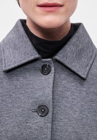 Casaco cinza com gola larga e botões pretos. O tecido parece macio com uma textura subtil, sobrepondo-se a uma blusa preta.