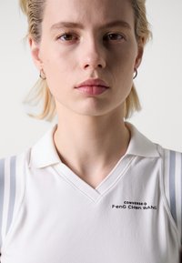 Polo shirt blanc avec col en V, rayures grises sur les épaules, et logo "CONVERSE Feng Chen Wang" imprimé sur la poitrine. Texture douce, design près du corps.