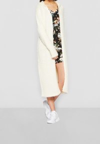 Lange, open-front cardigan van pluizig crèmekleurig materiaal, voorzien van een capuchon en een gestructureerde afwerking. Draagt over een bloemenjurk en witte sneakers.