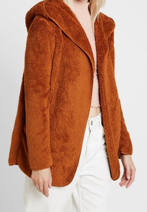 Roestkleurige, zachte jas met capuchon gedragen over een lichtroze geribbelde cropped top en witte broek met hoge taille.