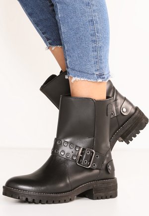 Bottes en cuir noires mi-mollet avec talons épais et sangles à boucle cloutées, portées avec un jean bleu clair raccourci et effiloché.