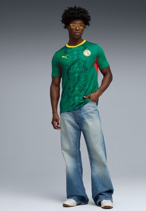 Moški, oblečen v zeleno nogometno majico z rumenim ovratnikom in rdečimi stranskimi paneli, svetlo modre široke kavbojke, bele superge ter rumeno obarvana očala.