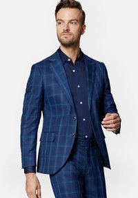 Granatowa marynarka w kratę z pasującymi spodniami, z dwoma guzikami, klapami oraz gładką fakturą. Pod spodem długa rękaw koszula.