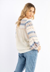 Blusa leggera di crema con strisce verticali blu e arancioni, maniche a sbuffo e tessuto testurizzato. Abbinata a jeans azzurri.