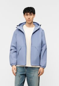 Νέος άνδρας με ανοιχτό μπλε μπουφάν με κουκούλα πάνω από λευκό ριμπ μπλουζάκι και μπλε τζιν, σε λευκό απλό φόντο.