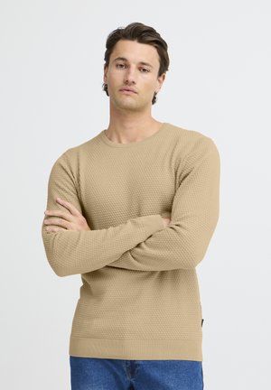 Suéter de textura beige con cuello redondo y puños de canalé, llevado sobre unos jeans azules. La tela presenta un sutil patrón de gofre.