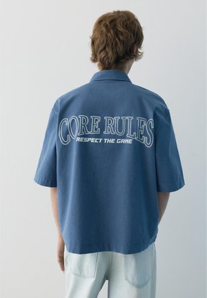 Persoană cu păr scurt și castaniu poartă o cămașă albastră largă cu textul „CORE RULES RESPECT THE GAME” pe spate, stând în fața unui fundal simplu.