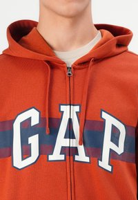 Crvena dukserica s patentnim zatvaračem sprijeda, s bijelim "GAP" logom i tamnoplavim prugama preko prsnog dijela. Meki materijal, podešive vrpce.