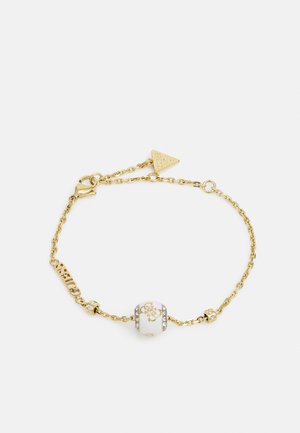 Bracelet en chaîne dorée avec une perle blanche ornée d'un motif gravé et d'accents en cristaux, terminé par un fermoir sécurisé et une étiquette en forme de triangle.