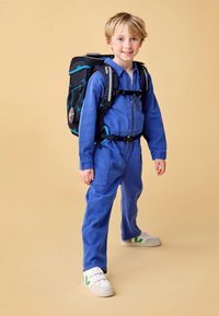 Jumpsuit zippé bleu avec des poignets élastiques et des poches, associé à un sac à dos noir et bleu. Baskets blanches avec des accents verts. Fond simple.