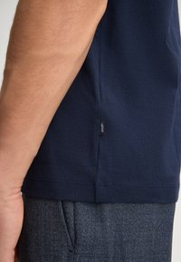 Marineblauwe katoenen T-shirt met een subtiel logo-label, korte mouwen en een zachte textuur. Gecombineerd met gestructureerde grijze broeken.