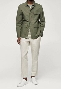 Giubbotto verde oliva con quattro tasche frontali, chiusura con bottoni, indossato sopra una camicia a righe. Abbinato a pantaloni beige e sneakers bianche.