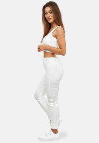 Mujer joven con cabello largo y marrón, vistiendo una camiseta corta blanca, jeans ajustados blancos y zapatillas blancas, de pie de lado y sonriendo.