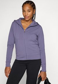 Violet zip-up kapucei ar kapuci, aprīkots ar sānu kabatām un ribu aprocēm, apvienots ar melniem legingiem. Gluda auduma tekstūra.