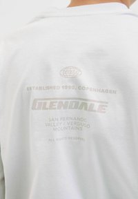 Balta kapuce ar grafisku tekstu uz muguras, kas ietver "GLENDALE", atrašanās vietas datus un "ESTABLISHED 1990" maigos toņos.
