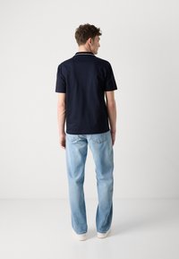 Camiseta polo navy con acentos de cuello blanco, mangas cortas, combinada con jeans de color azul claro y zapatillas blancas, vista desde atrás.