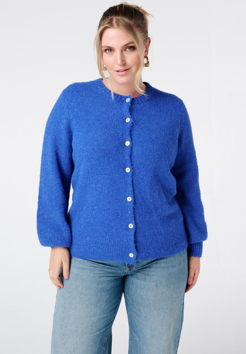 Blauwe gebreide cardigan met een ronde hals, knoopsluiting aan de voorkant, lange mouwen en een zachte textuur. Voorzien van witte knopen. Draagt over lichtblauwe spijkerbroek.