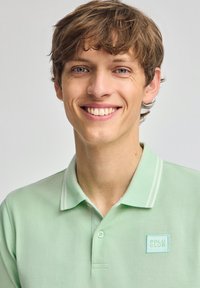 Jasnozielona koszulka polo z teksturowanej tkaniny, z dwiema białymi paskami na kołnierzyku oraz małym haftowanym logo "POLO CLUB" na piersi.