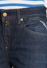Jeans in denim blu scuro con cuciture arancioni, dotati di bottoni e rivetti in metallo, e un piccolo etichetta del marchio vicino alla tasca.