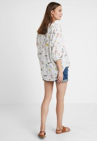 Blusa branca com padrão colorido de praia, de corte solto e com barra arredondada. Combinada com calções de ganga e sandálias castanhas.