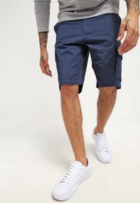 Shorts cargo bleu marine en mélange de coton, dotés de poches latérales, d'une coupe droite et d'une texture lisse. Associés à des baskets blanches.