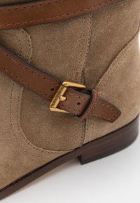 Bottine en suède beige avec une bride en cuir marron dotée d'une boucle dorée. Couture remarquable et talon en bois lisse.