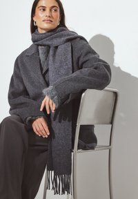 Manteau en laine gris foncé avec une texture duveteuse, accompagné d'une écharpe assortie avec des bords à franges. Modèle assis, les mains reposant sur une chaise en métal.
