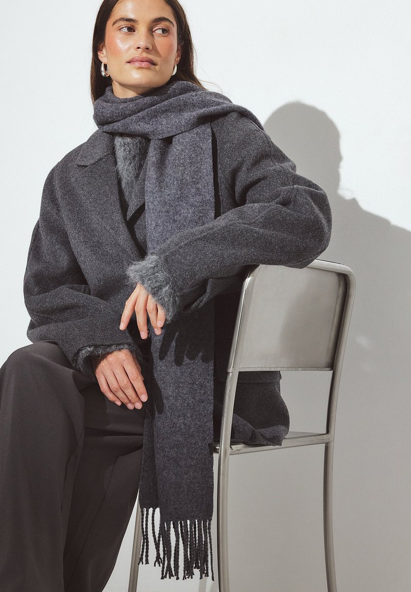 Manteau en laine gris foncé avec une texture duveteuse, accompagné d'une écharpe assortie avec des bords à franges. Modèle assis, les mains reposant sur une chaise en métal.
