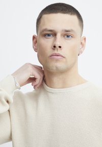 Pulover tricotat bej cu guler rotund; modelul are părul scurt și își atinge gâtul. Brățară argintie pe încheietura stângă. Ochii albaștri.