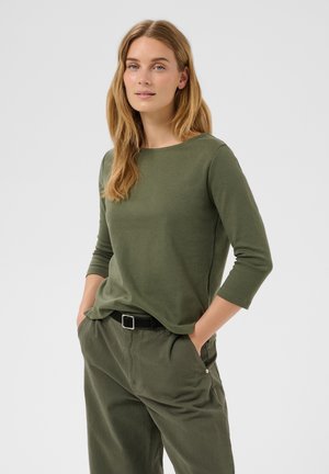Młoda kobieta o długich blond włosach, ubrana w dopasowaną zieloną koszulę z rękawem do łokcia i spodnie z czarnym paskiem, ręce w kieszeniach, neutralne tło.