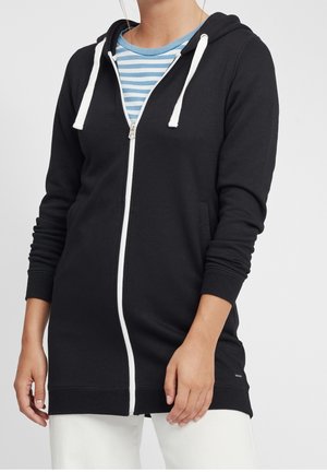 Femme portant un long sweat à capuche noir zippé avec des cordons blancs par-dessus une chemise rayée bleue et blanche et un pantalon blanc.