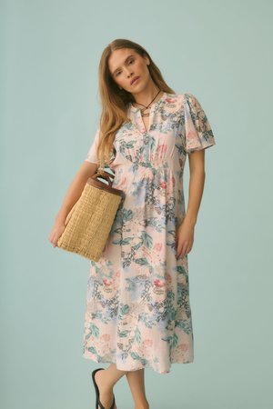 Vrouw draagt een pastelgekleurde bloemenmidi-jurk met pofmouwen en houdt een gevlochten strohandtas vast tegen een effen lichtblauwe achtergrond.