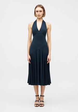 Γυναίκα με μίντι φόρεμα μπλε ναυτικού χρώματος χωρίς μανίκια και με λαιμόκοψη halter, φούστα με πτυχώσεις και μαύρα ψηλοτάκουνα σανδάλια με λουριά, στέκεται μπροστά σε απλό φόντο.