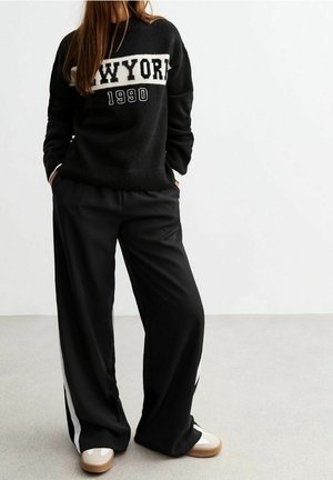 Personne portant un pull noir New York 1990, un pantalon noir ample avec des bandes blanches sur les côtés et des baskets beiges, debout sur un fond blanc uni.