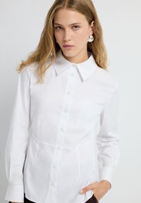 Giovane donna con capelli castano chiaro che indossa una camicia bianca con colletto e grandi orecchini di argento, posando davanti a uno sfondo bianco uniforme.