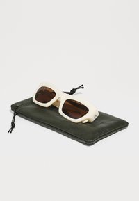 Des lunettes de soleil avec des montures crème et des verres foncés, reposant sur une pochette textured olive verte avec une fermeture à cordon.
