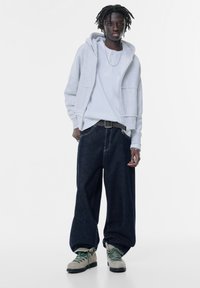Grå huvtröja över en vit skjorta, tillsammans med lösa mörka denimjeans och grå stövlar med gröna snören. Enkel design, avslappnad passform.