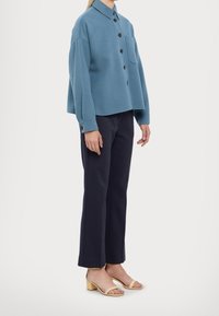 Giacca button-up blu chiaro con tasca sul petto, abbinata a pantaloni blu navy e sandali gialli con tacco a blocco. Tessuto morbido, vestibilità rilassata.