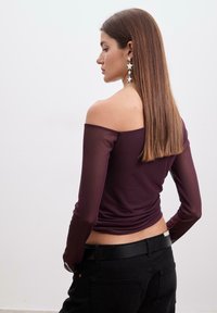Burgundy off-shoulder top med lange, gennemsigtige ærmer, kombineret med sorte højblussede bukser og et sort bælte, der fremhæver en glat tekstur.