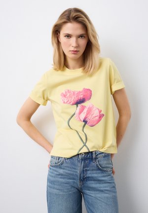 Giovane donna che indossa una maglietta gialla con grafica di papaveri rosa e jeans blu, in piedi con le mani sui fianchi contro uno sfondo bianco.