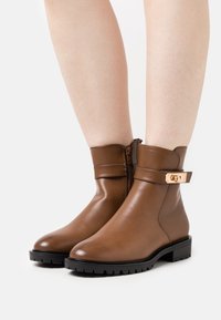 Bottines en cuir marron avec une finition lisse et une fermeture éclair sur le côté. Elles disposent d'une sangle décorative avec une boucle en métal doré. Semelles en caoutchouc noir.
