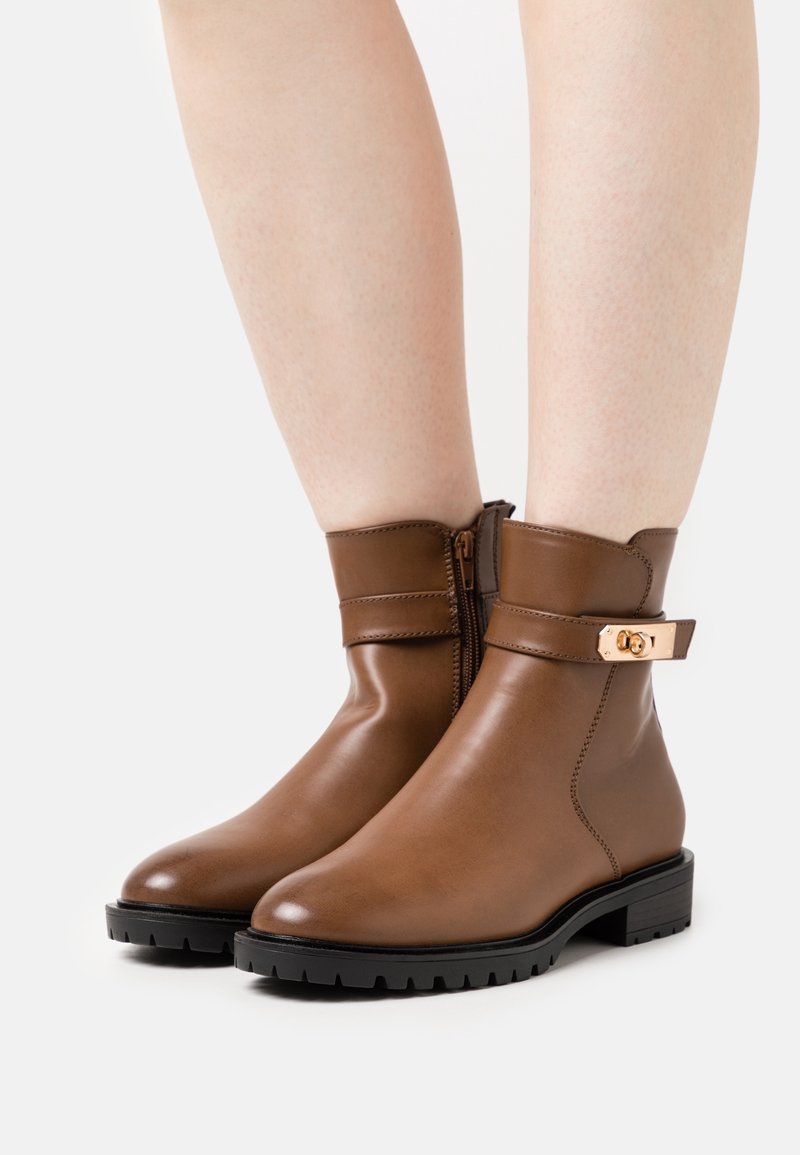 Bottines en cuir marron avec une finition lisse et une fermeture éclair sur le côté. Elles disposent d'une sangle décorative avec une boucle en métal doré. Semelles en caoutchouc noir.