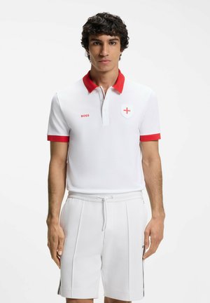 Hombre vestido con una camiseta polo blanca con cuello y puños rojos, un emblema de escudo de cruz roja y pantalones cortos blancos con cordón, de pie frente a un fondo sencillo.