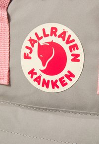 Šedá tkanina s ružovými popruhmi obsahuje kruhovú nášivku so zobrazením loga červenej líšky a textom "FJÄLLRÄVEN KÅNKEN" písmom tučným.